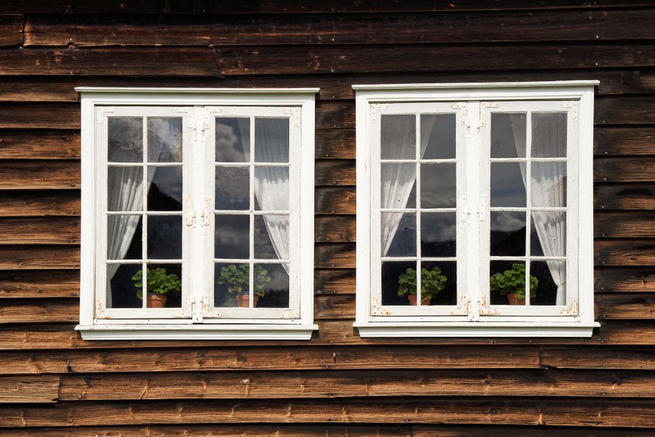 Two white wood-framed casement windows with curtains and potted plants on a dark wood plank wall