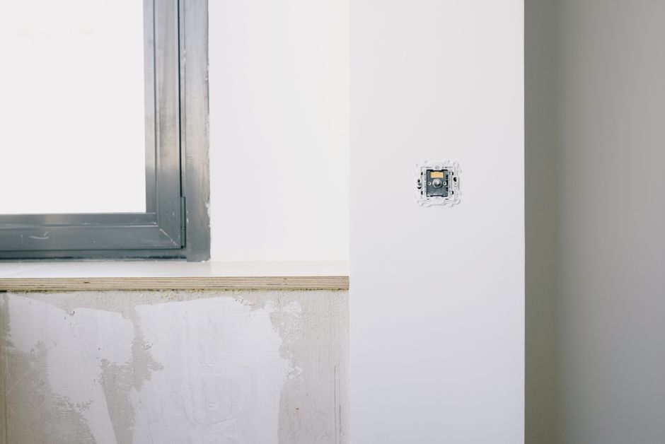 Window in a dark metal frame beside a plywood sill and a wall switch plate in an unfinished room