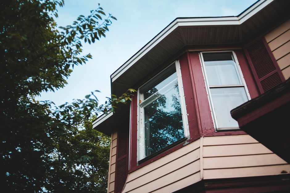 Corner of a house with a white-framed window and a dark red painted bay under green trees