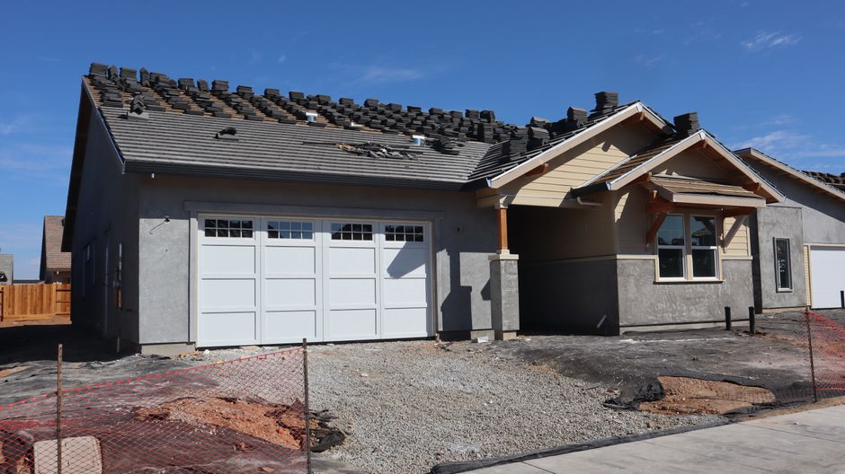 Stucco house under construction with a garage door, a tile roof being laid and a window beside the entry