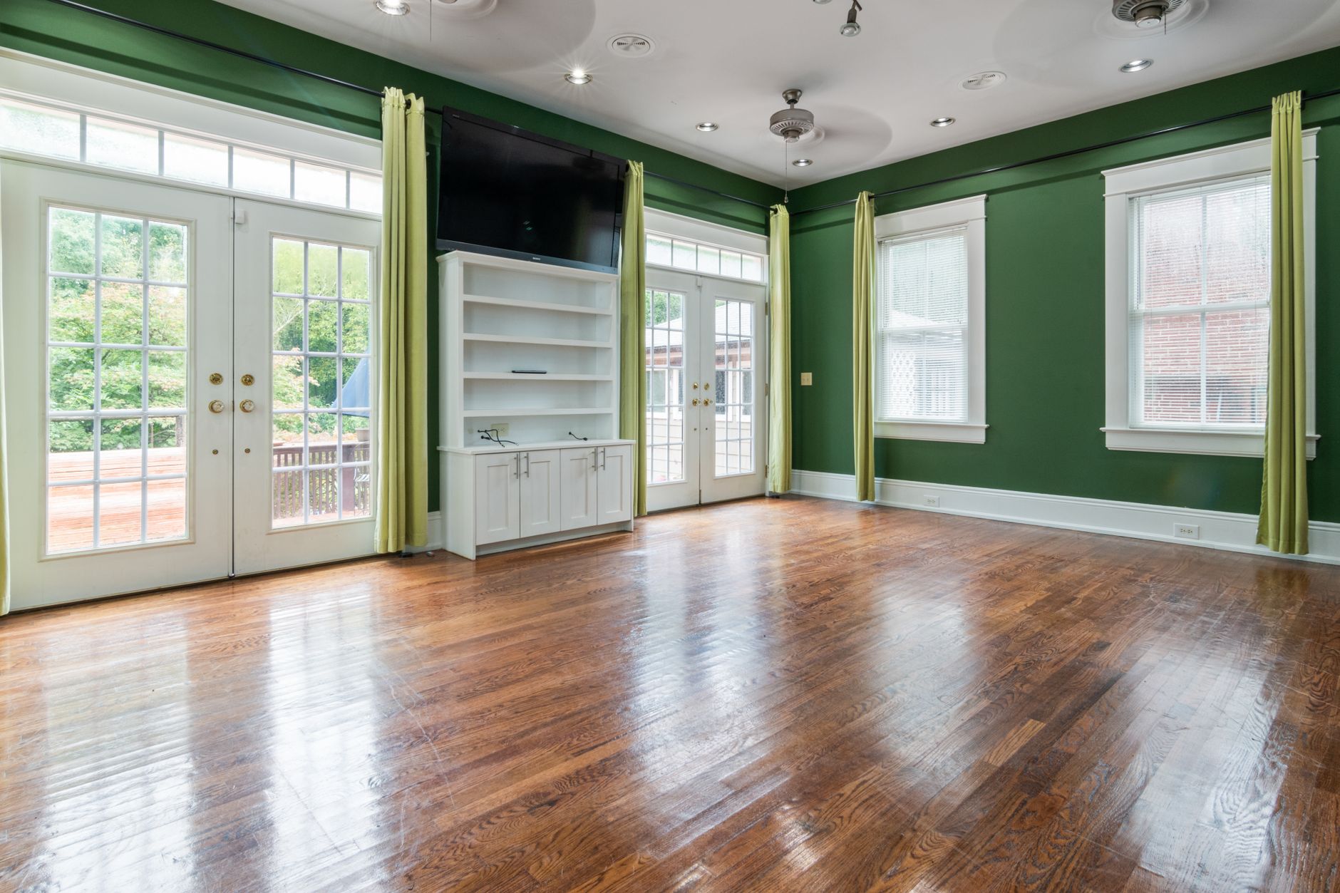 Empty sunlit room with a hardwood floor, large windows and green walls